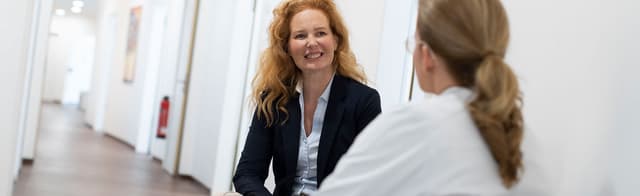 Dieses Bild zeigt Frau Dr. Jennifer Quist im Gespräch mit einer Arbeitskollegin auf dem Gang in der Praxis.