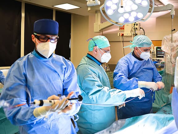 Ärzteteam im Operationssaal in blauen Schutzkitteln und Masken führt einen Eingriff durch. Ein Arzt hält ein katheterähnliches Instrument, während zwei Kolleg:innen am Operationstisch arbeiten. Helles OP-Licht beleuchtet den Bereich; der Patient liegt verdeckt unter Tüchern.