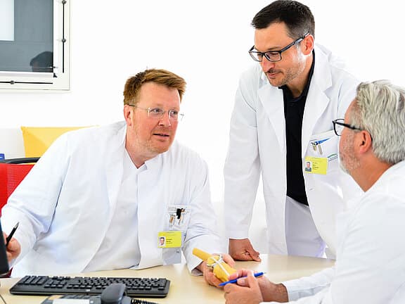 Drei Ärzte in weißen Kitteln besprechen am Schreibtisch einen Fall; einer hält ein Herzmodell, ein Computer liegt vor ihnen. Teamarbeit in einer Klinik.