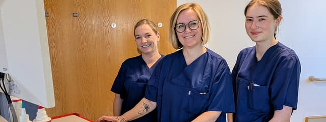 Drei Personen in blauen Pflegeuniformen stehen lächelnd an einem Computerarbeitsplatz in einer Klinik und blicken zur Kamera; auf dem Tisch liegen medizinische Utensilien.