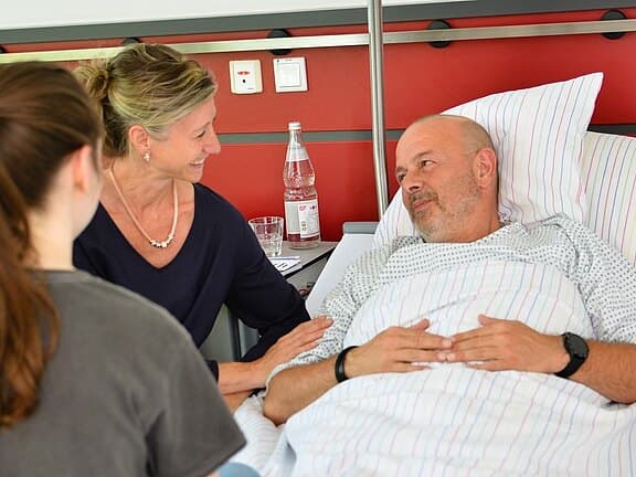 Krankenhauszimmer: Ein männlicher Patient liegt im Bett, blickt lächelnd zu einer Frau, die seine Hand berührt. Neben ihr sitzt eine weitere Person. Die Szene wirkt fürsorglich und ruhig, zeigt einen unterstützenden Besuch am Krankenbett.