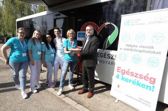 A szűrőprogram csapata: Dr. Laczkó Tamás főigazgató öltönyben, mellette öt fiatal egészségügyi szakdolgozó türkizkék pólóban és fehér nadrágban áll a szűrőbusz és az "Egészség 4 keréken!" feliratú molinó előtt.