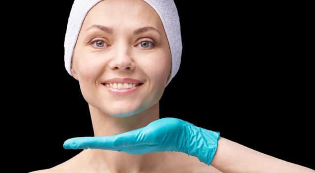 Smiling woman in spa headband getting a skincare assessment at a dermatology clinic, examined by a professional in blue gloves.