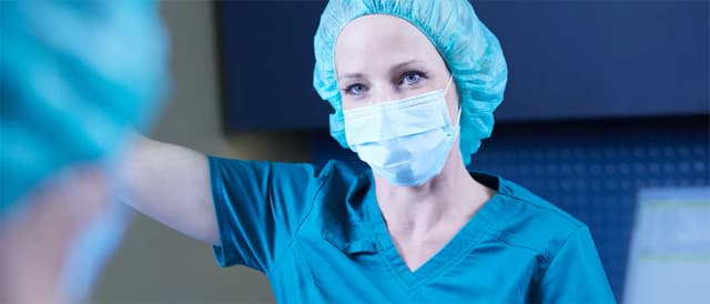 Chirurgin in blauer OP-Kleidung mit Maske und Haube, konzentriert während eines medizinischen Eingriffs