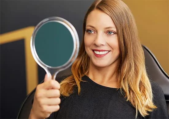 Eine Frau mit blondem Haar lächelt in einen Handspiegel, im Hintergrund dunkle und goldene Töne