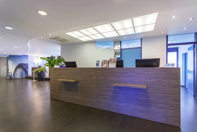 Moderner Empfangsbereich der Klinik am Ring in Köln mit großer Holz- und Glas-Front, zwei Computerarbeitsplätzen, Infomaterial und Pflanzen auf der Theke sowie heller Deckenbeleuchtung.
