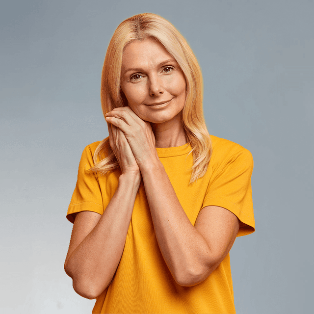 Eine lächelnde Frau mittleren Alters mit blonder Schulterfrisur steht vor einem gelben Hintergrund und hält ihre Hände sanft an die Wange – Symbol für Wohlbefinden, Hautgesundheit und Anti-Aging.