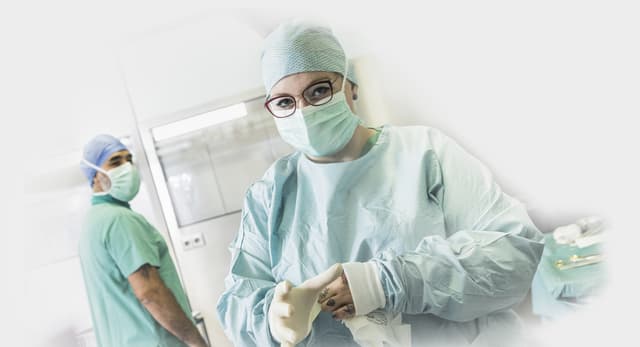 Junge Ärztin mit Brille, Mundschutz, Haarnetz und OP-Kittel zieht sich Handschuhe an.