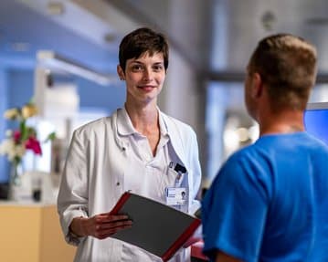 Eine Krankenschwester in einem weißen Kittel hält eine Akte und spricht mit einem Patienten in einem blauen T-Shirt. Der Hintergrund zeigt einen Krankenhausflur mit Blumen und medizinischen Geräten.