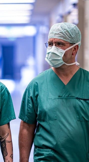 Drei medizinische Fachkräfte in grünen OP-Kitteln und Mundschutz gehen durch einen Krankenhausflur. Sie tragen Operationsmützen und haben einen konzentrierten Ausdruck.