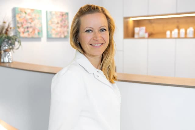 Frau in weißem Kittel vor moderner Inneneinrichtung mit Kunstwerken und Regalen im Hintergrund.