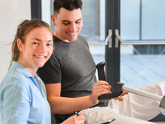 Physiotherapeutin begleitet einen jungen Mann beim Training an einem Ergometer; beide lächeln. Die Therapeutin notiert Werte auf einem Klemmbrett, der Patient hält den Griff des Geräts. Szene vermittelt Reha oder gesundheitsorientiertes Training in heller Umgebung.